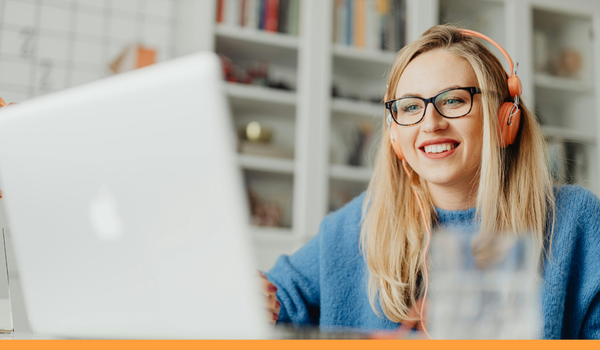 A woman with long blonde hair and glasses smiling while working on a laptop. She is wearing a blue jumper and orange headphones.