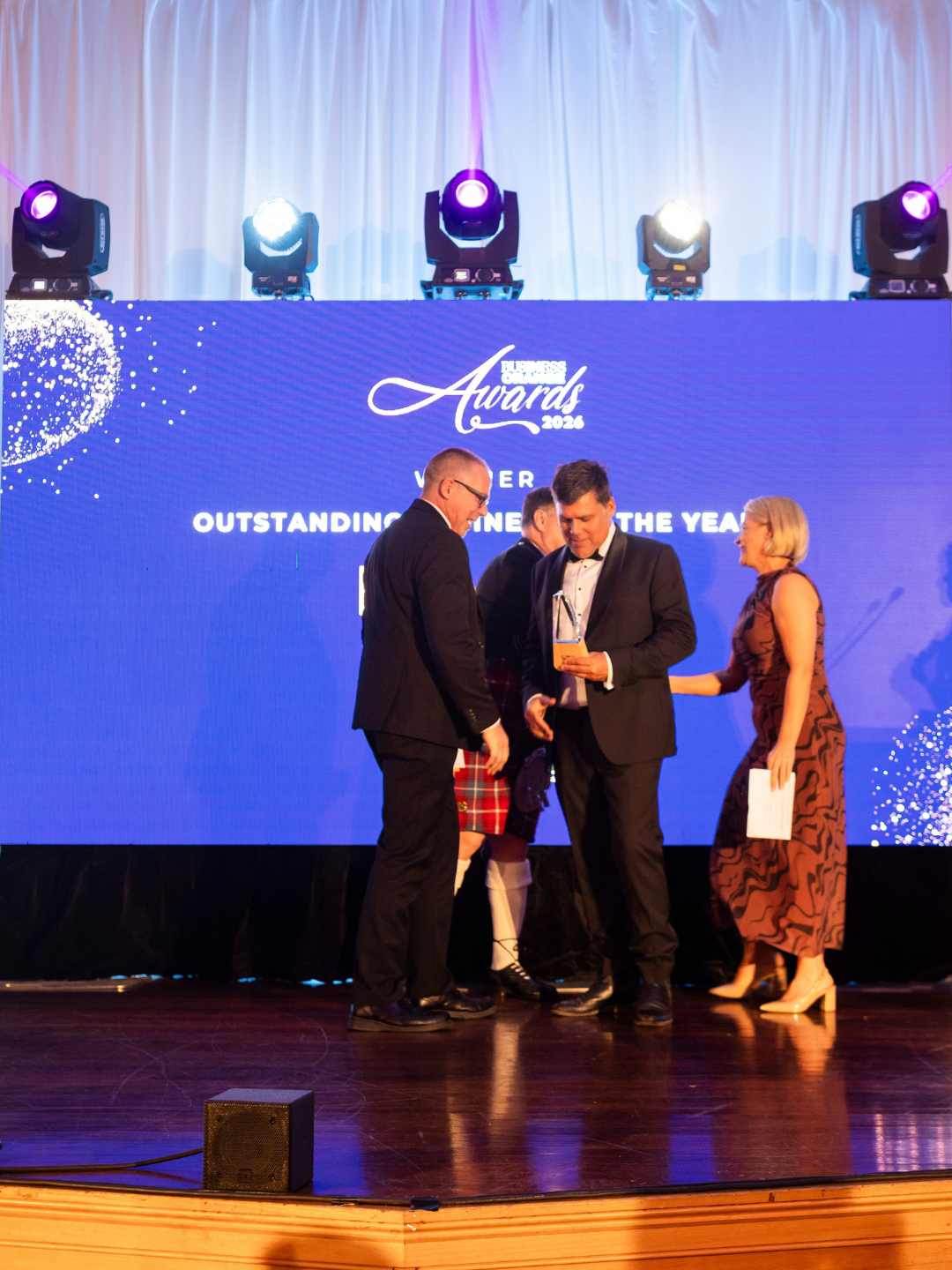 winners accepting trophy on stage in front of large indigo screen